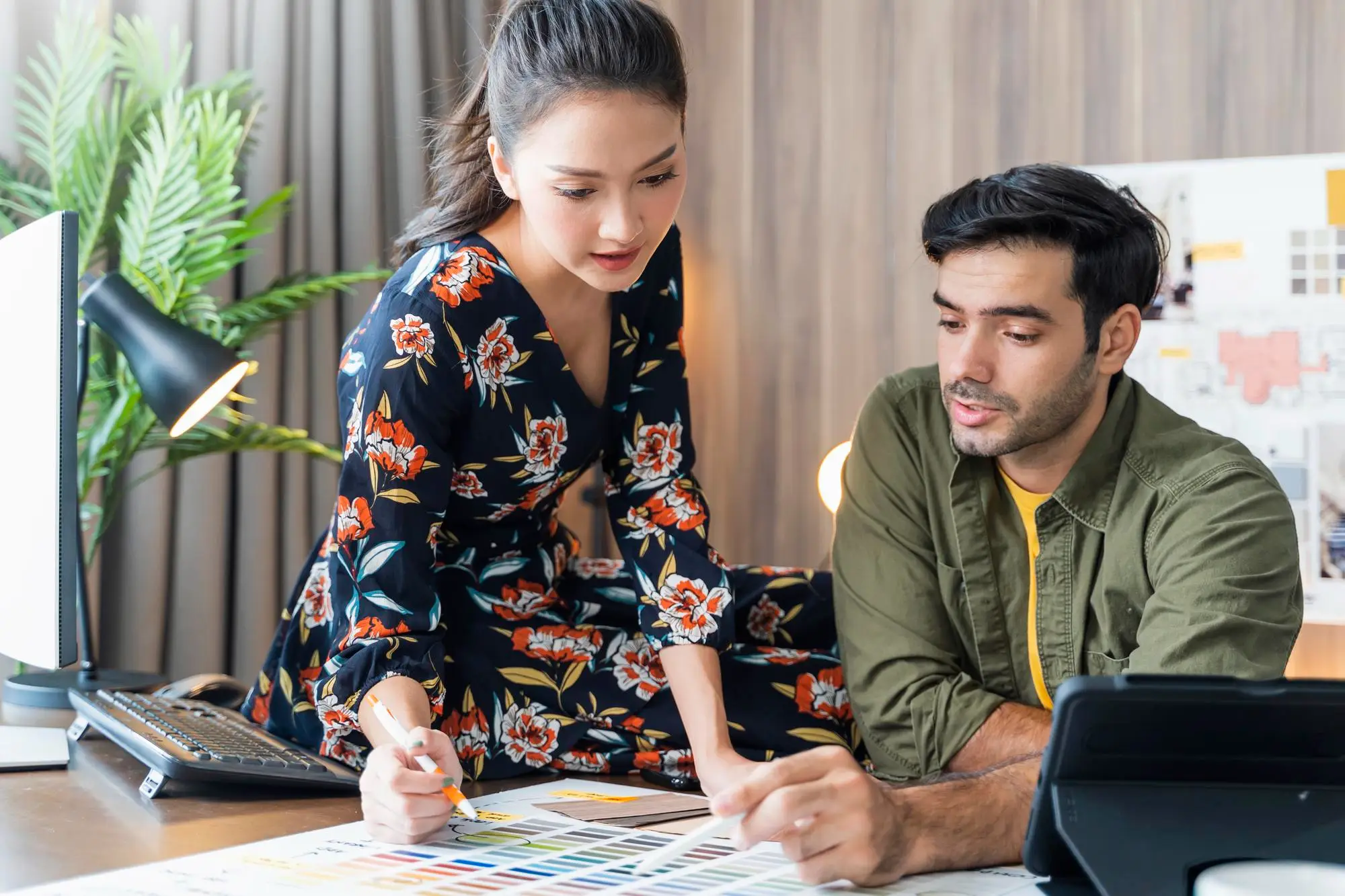 A woman in a floral dress and a man in a green shirt review color swatches together at a desk with a computer, tablet, and paperwork in a modern office setting.