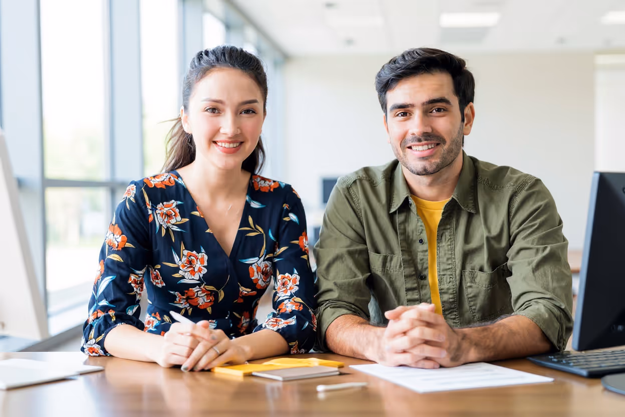 A smiling woman in a floral blouse and a man in a green shirt sit side by side at a desk in a bright office, facing the camera with papers and a computer nearby.