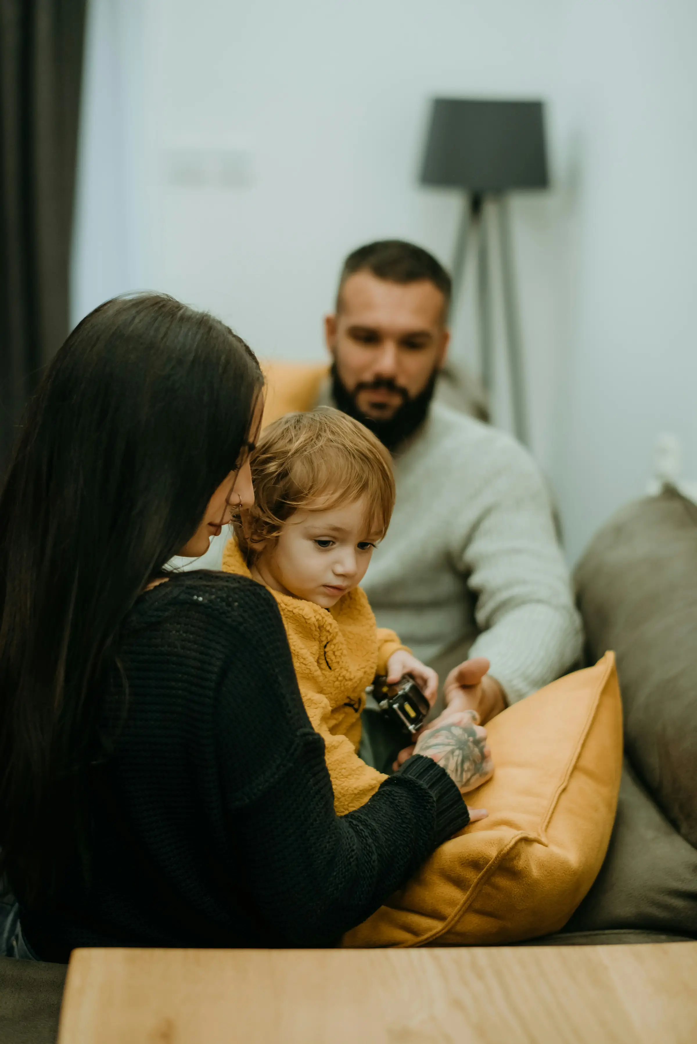 A woman holds a young child on her lap while sitting on a couch. A man sits next to them, watching. They are indoors in a cozy, softly lit living room with cushions and a floor lamp.