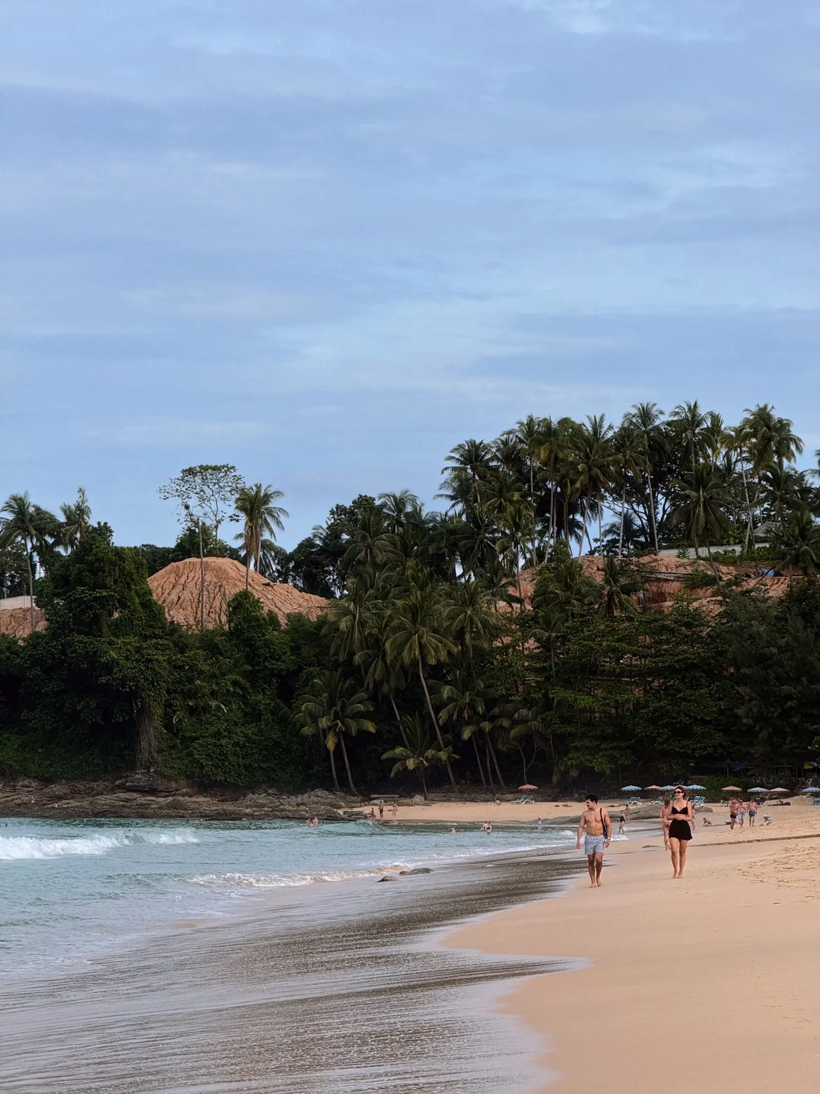 Tropical beach in Phuket, Thailand, with palm trees, gentle waves, sandy shoreline, and visitors walking by the sea.