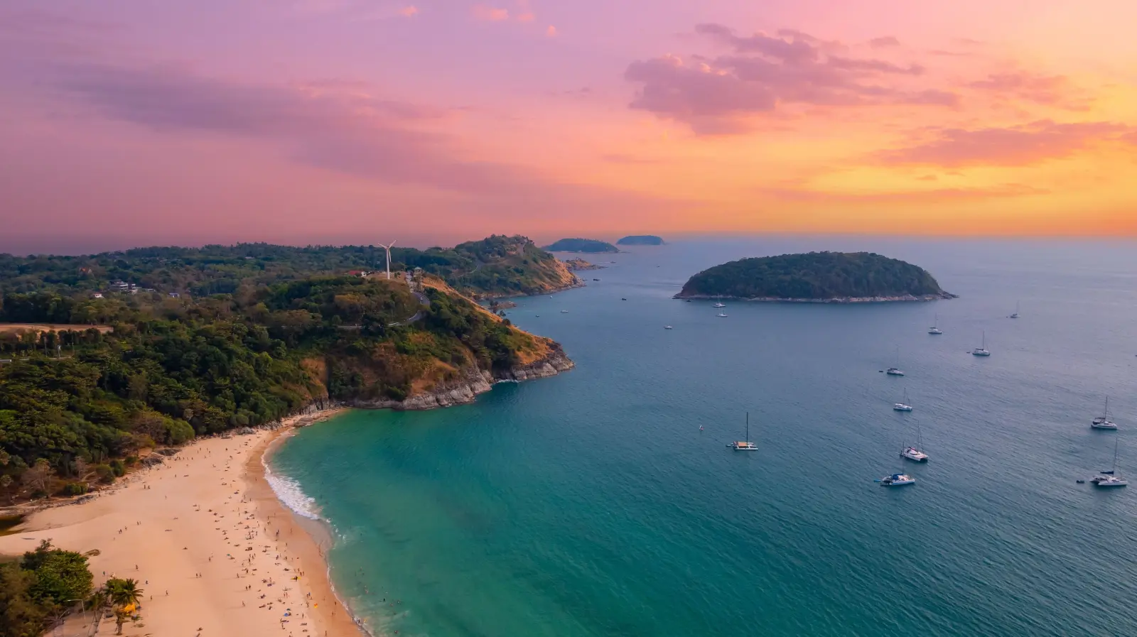  Aerial view of Nai Harn Beach in Phuket, Thailand, at sunset with turquoise water, anchored sailboats, and a sandy tropical coastline.