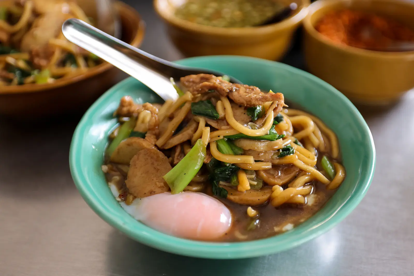 Close-up of Phuket-style Hokkien noodles with pork, vegetables, fish balls, and a soft-boiled egg in a savory sauce.