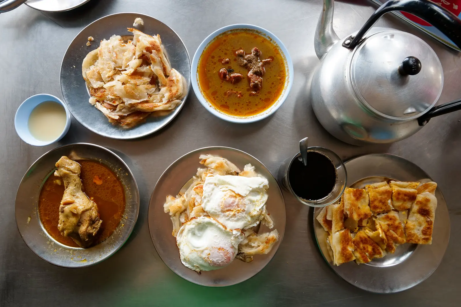 Phuket-style roti served with curry, fried eggs, chicken, dipping sauces, and hot tea on a stainless steel table.