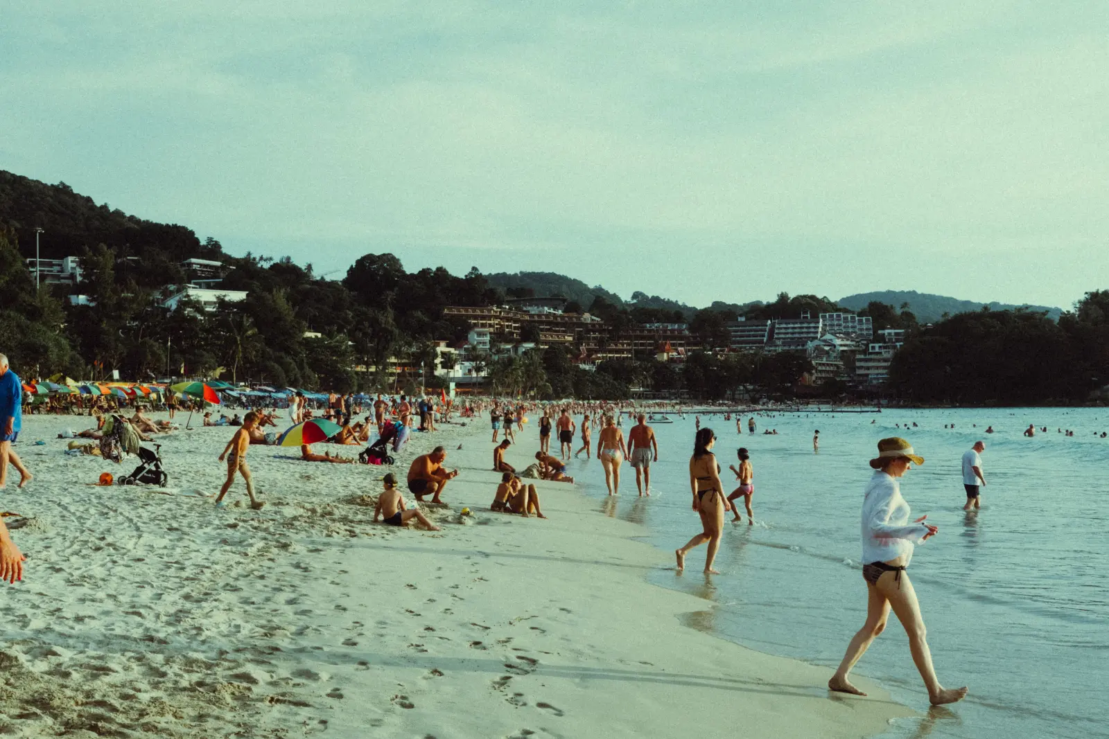 Kata Beach in Phuket, Thailand, with visitors relaxing on the sandy shoreline and swimming in calm tropical water.