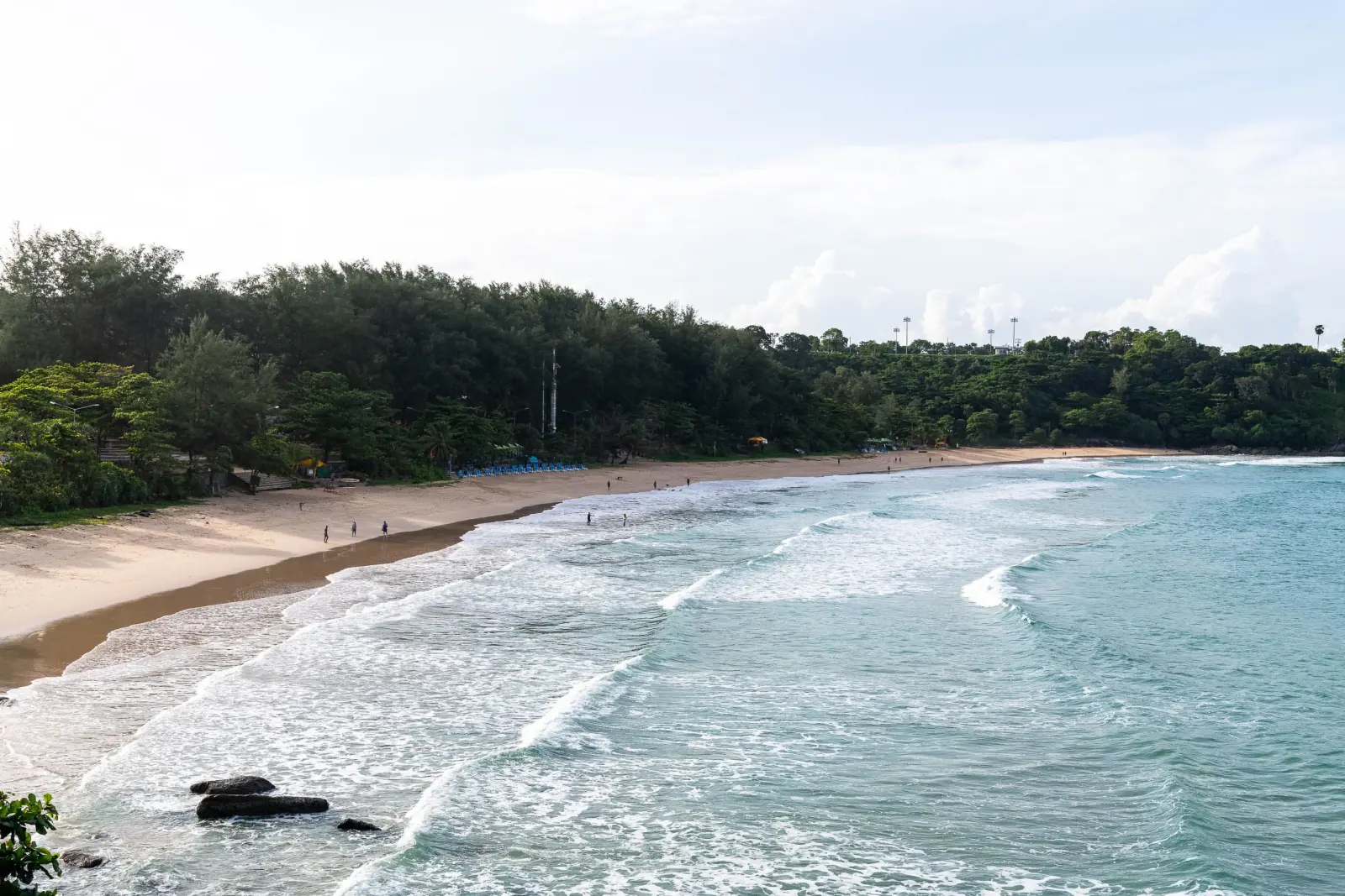 Nai Harn Beach in Phuket, Thailand, with a wide sandy shoreline, gentle waves, and lush green hills.