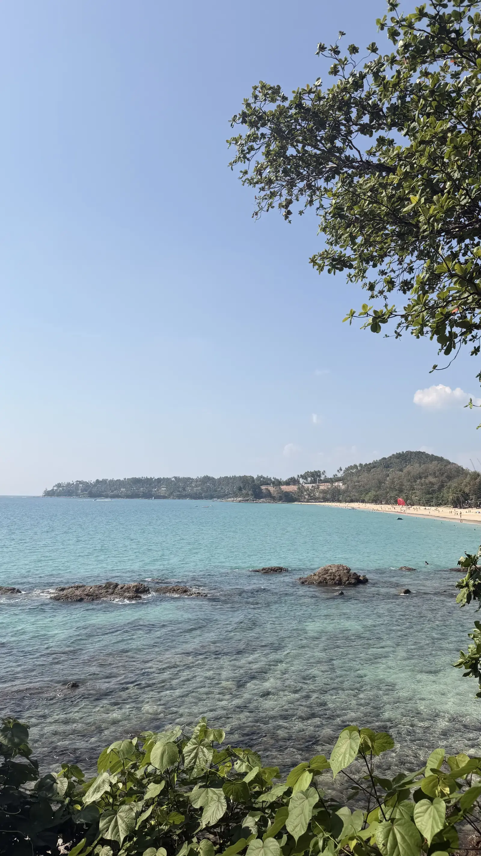 Surin Beach in Phuket, Thailand, with clear turquoise water, rocky shoreline, sandy beach, and tropical greenery.