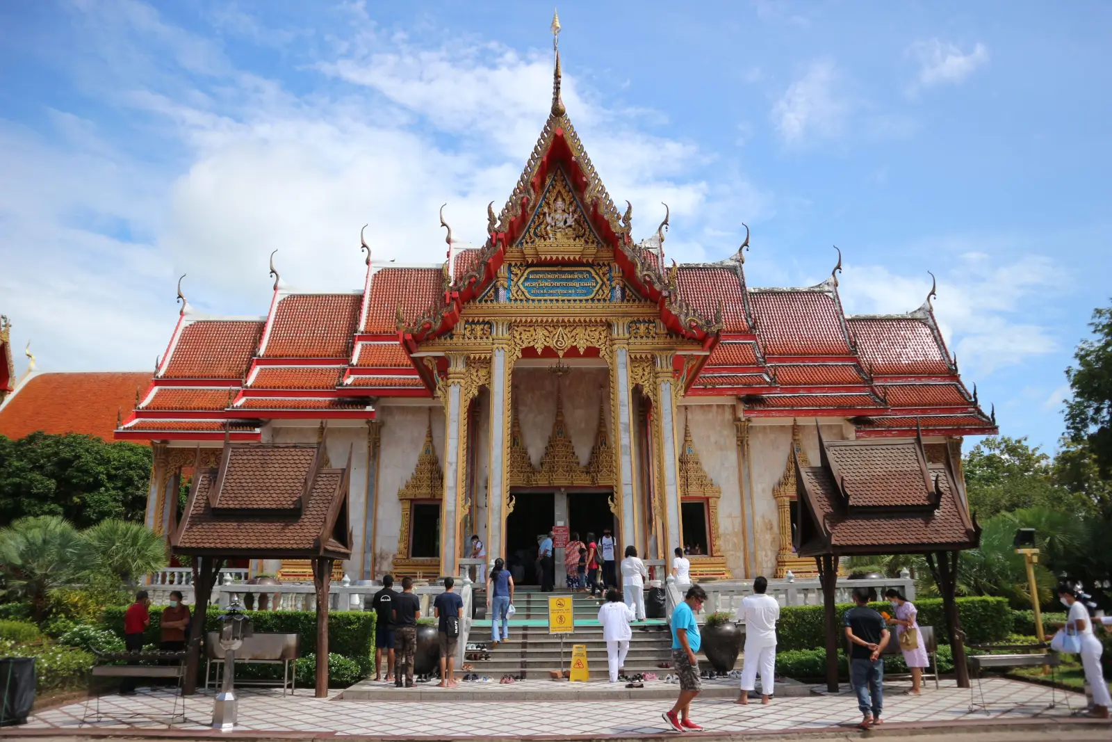 Wat Chalong temple in Phuket, Thailand, with ornate Thai architecture, red tiled roofs, and visitors entering the main hall.