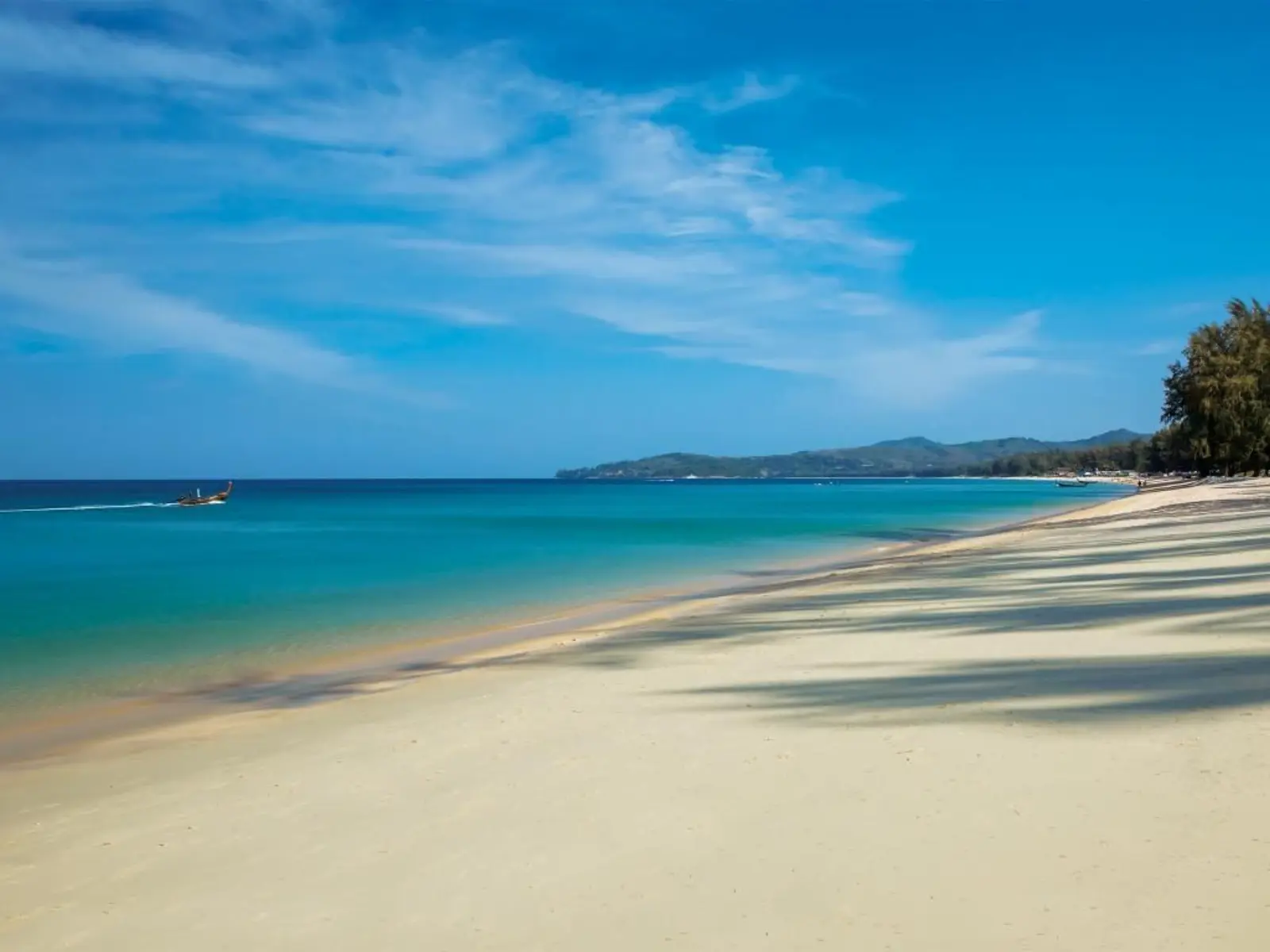 Bang Tao Beach in Phuket, Thailand, with a long sandy shoreline, clear turquoise water, and a traditional longtail boat offshore.