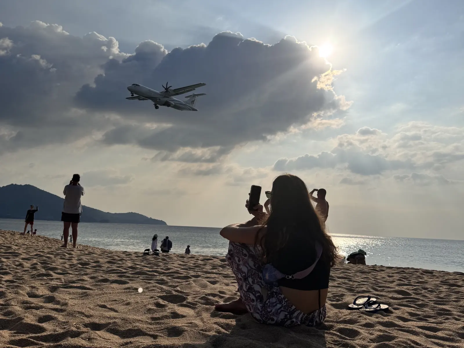 Plane flying low over Mai Khao Beach near Phuket International Airport as visitors watch and take photos from the sand.