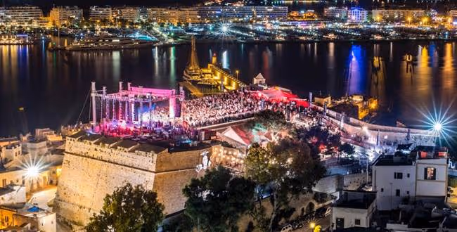 A large nighttime electronic music event set against a coastal cityscape. The stage is brightly lit with pink and purple lights, surrounded by a dense crowd. The venue is a historic fort overlooking a harbor filled with yachts and illuminated buildings, creating a vibrant seaside atmosphere.