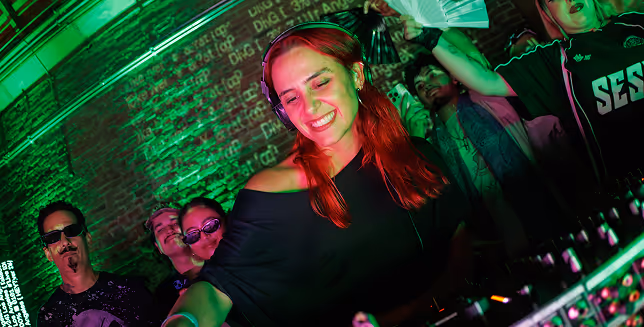 A DJ with red hair smiles while performing at a club, surrounded by people dancing and cheering under green lighting against a brick wall backdrop.