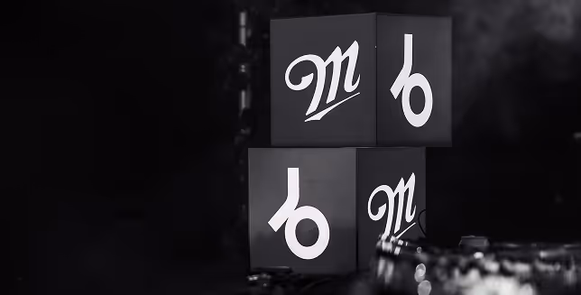 Black and white cubes stacked together, each featuring a stylized lowercase “b” and “m” logo, placed on a dark surface with a smoky background.