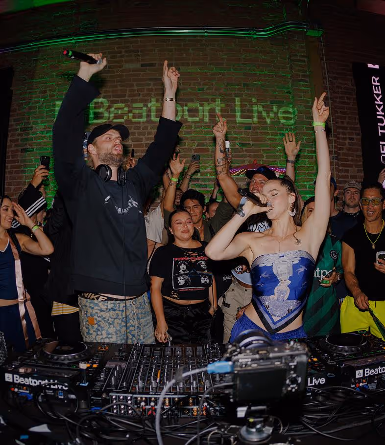 A lively DJ performance in a packed club, with two performers on stage—one holding a microphone and singing passionately, the other with headphones raising their hands as the crowd cheers. Colorful lights and a ‘Beatport Live’ sign glow on the brick wall behind them.