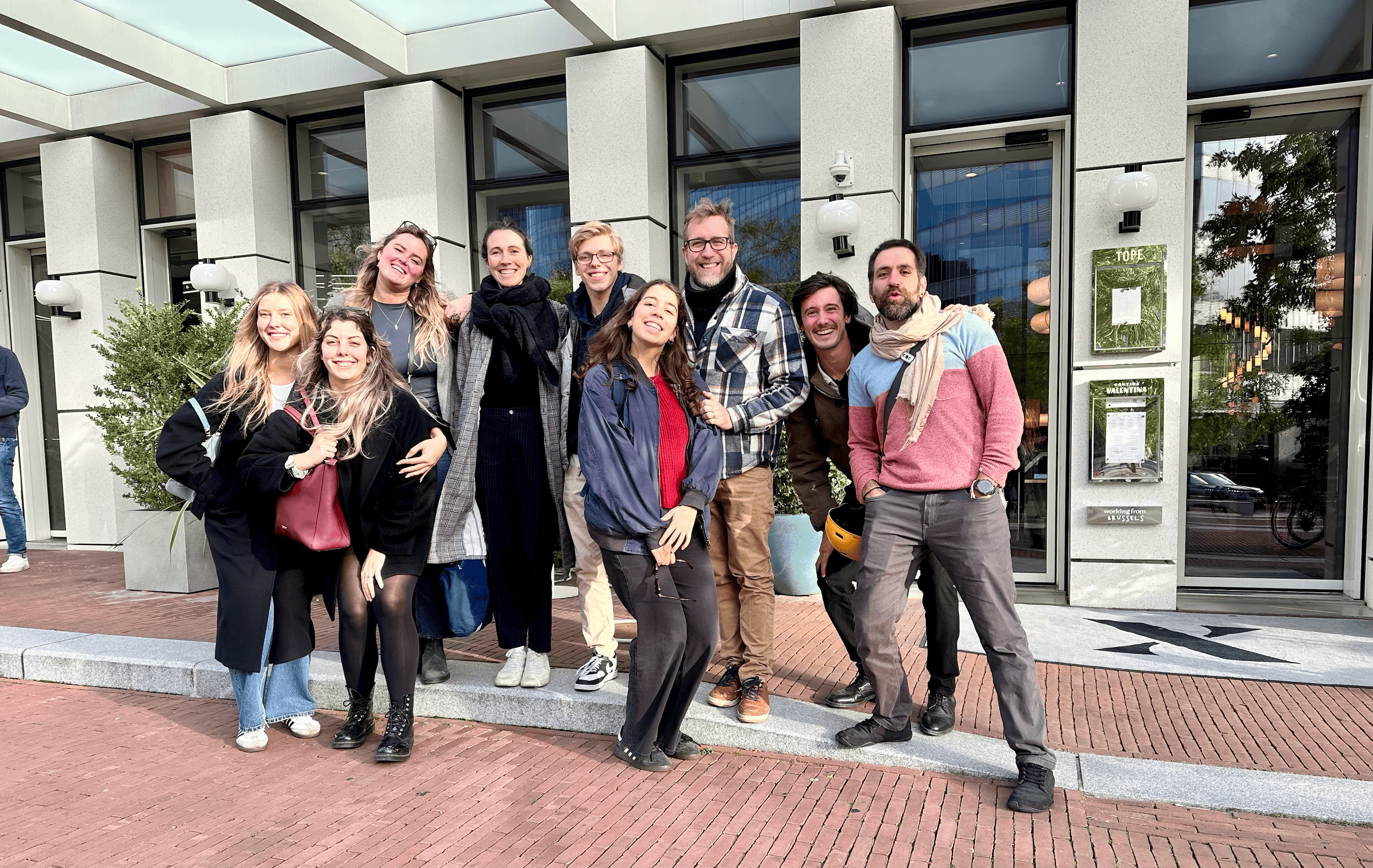 Group of nine smiling people standing outside a building with large windows, posing together on a sidewalk.