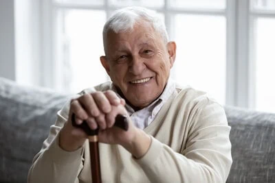 Smiling elderly man with white hair sitting indoors holding a wooden cane.