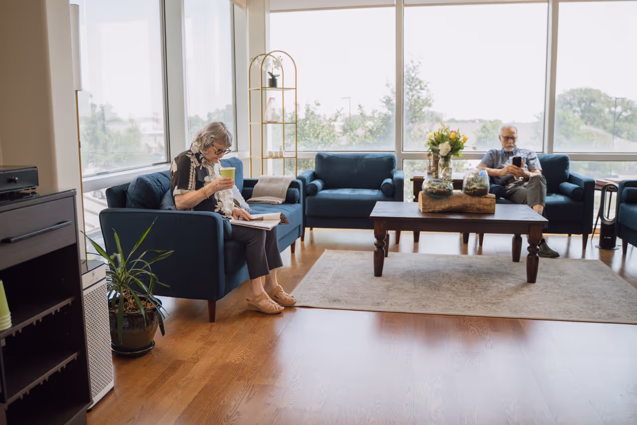 Elderly woman sitting on a blue sofa drinking from a green cup and reading a book in a bright living room with large windows, while an elderly man sits on another blue sofa using a phone.