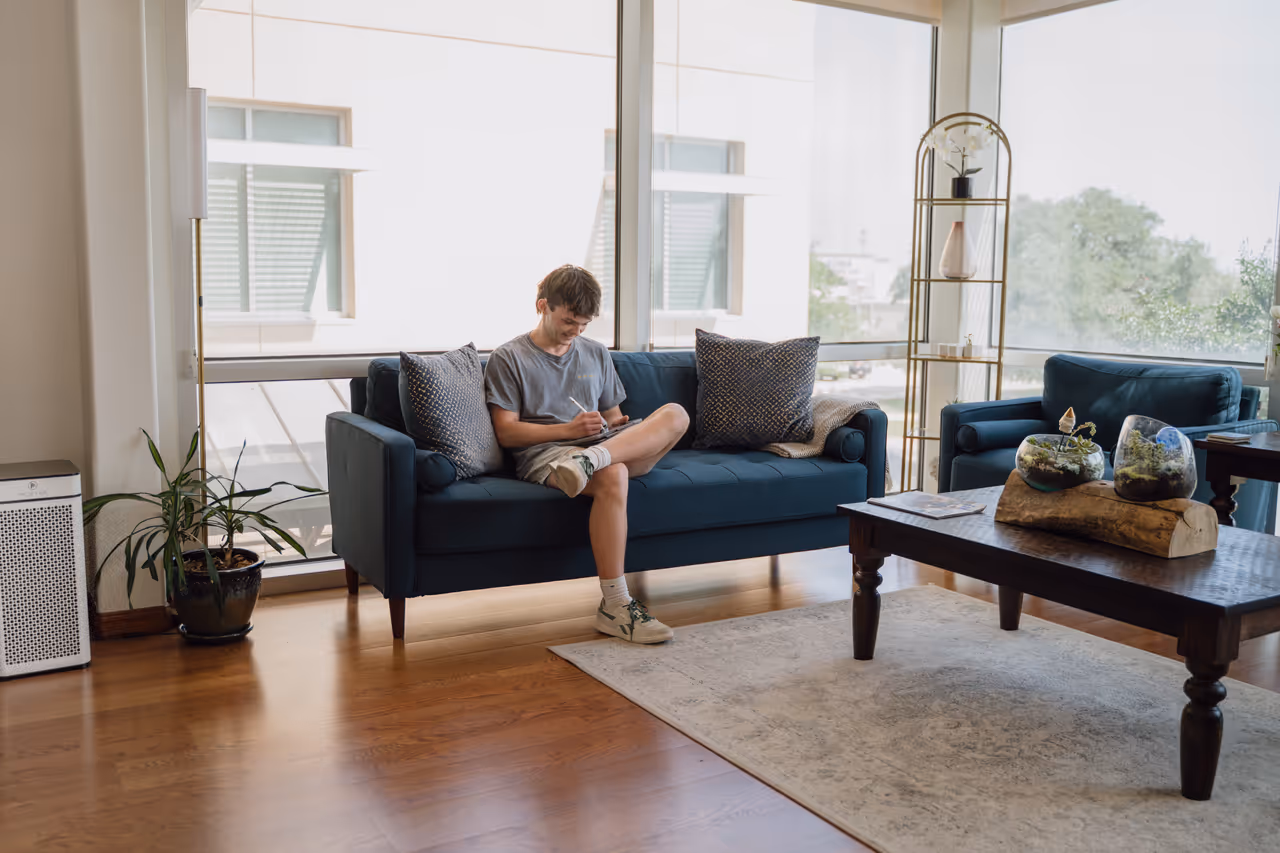 Young man sitting on a blue sofa writing in a notebook in a bright living room with large windows.
