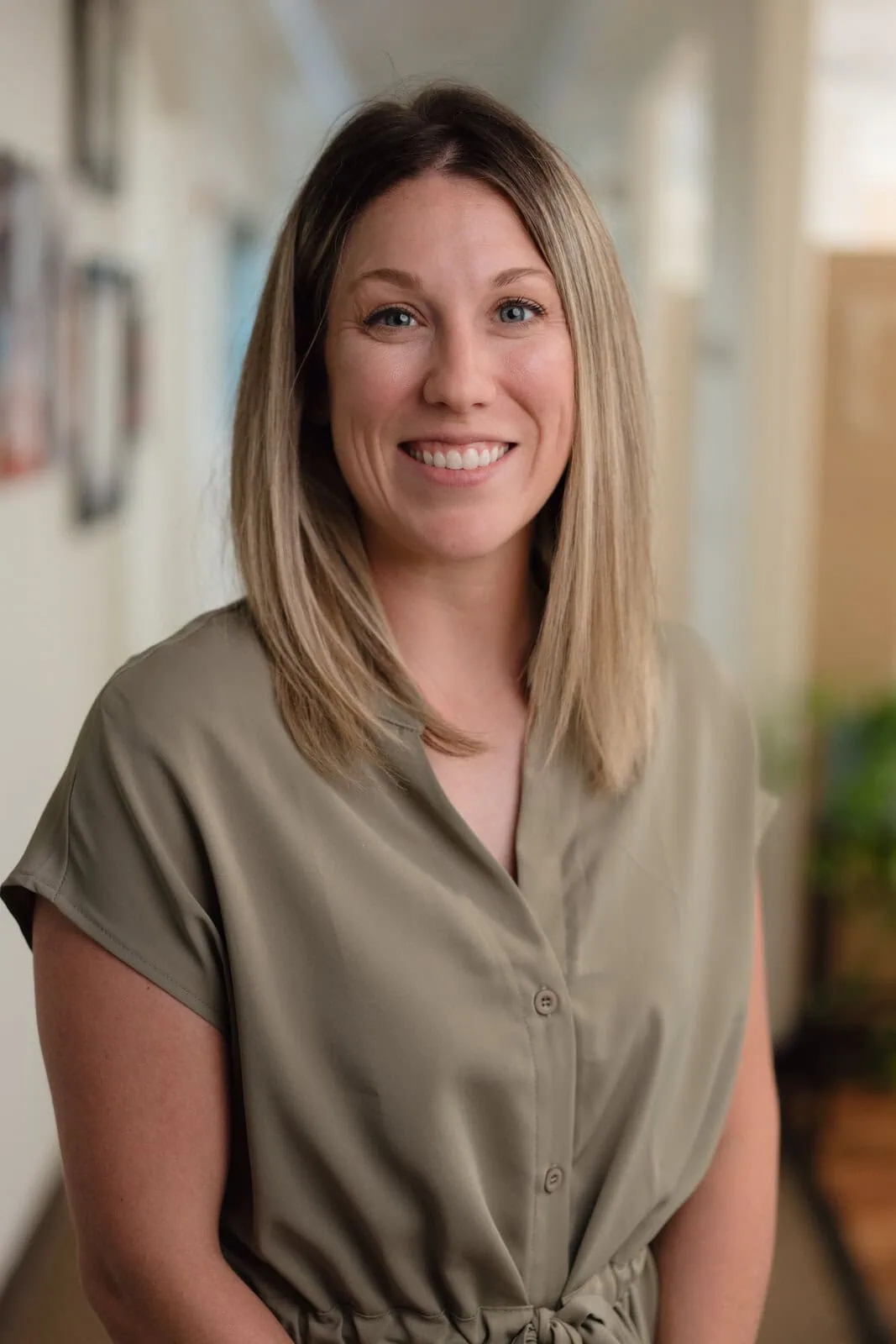 Smiling woman with shoulder-length blonde hair wearing a beige short-sleeve button-up dress in a softly lit indoor setting.