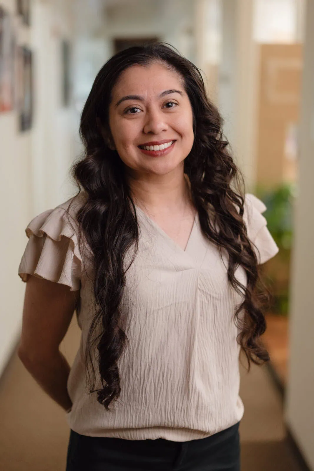 Smiling woman with long curly dark hair wearing a beige blouse standing in a softly lit hallway.
