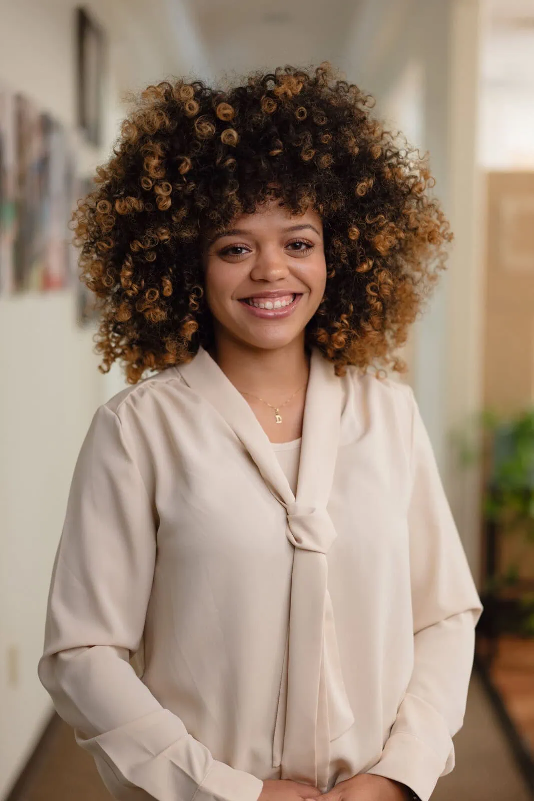 Smiling woman with voluminous curly hair wearing a beige blouse standing in a softly lit hallway.