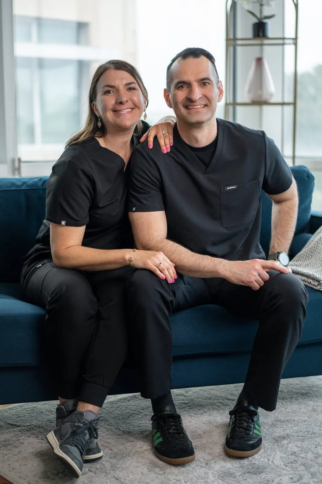 Smiling man and woman wearing black scrubs sitting closely on a blue couch in a bright room.