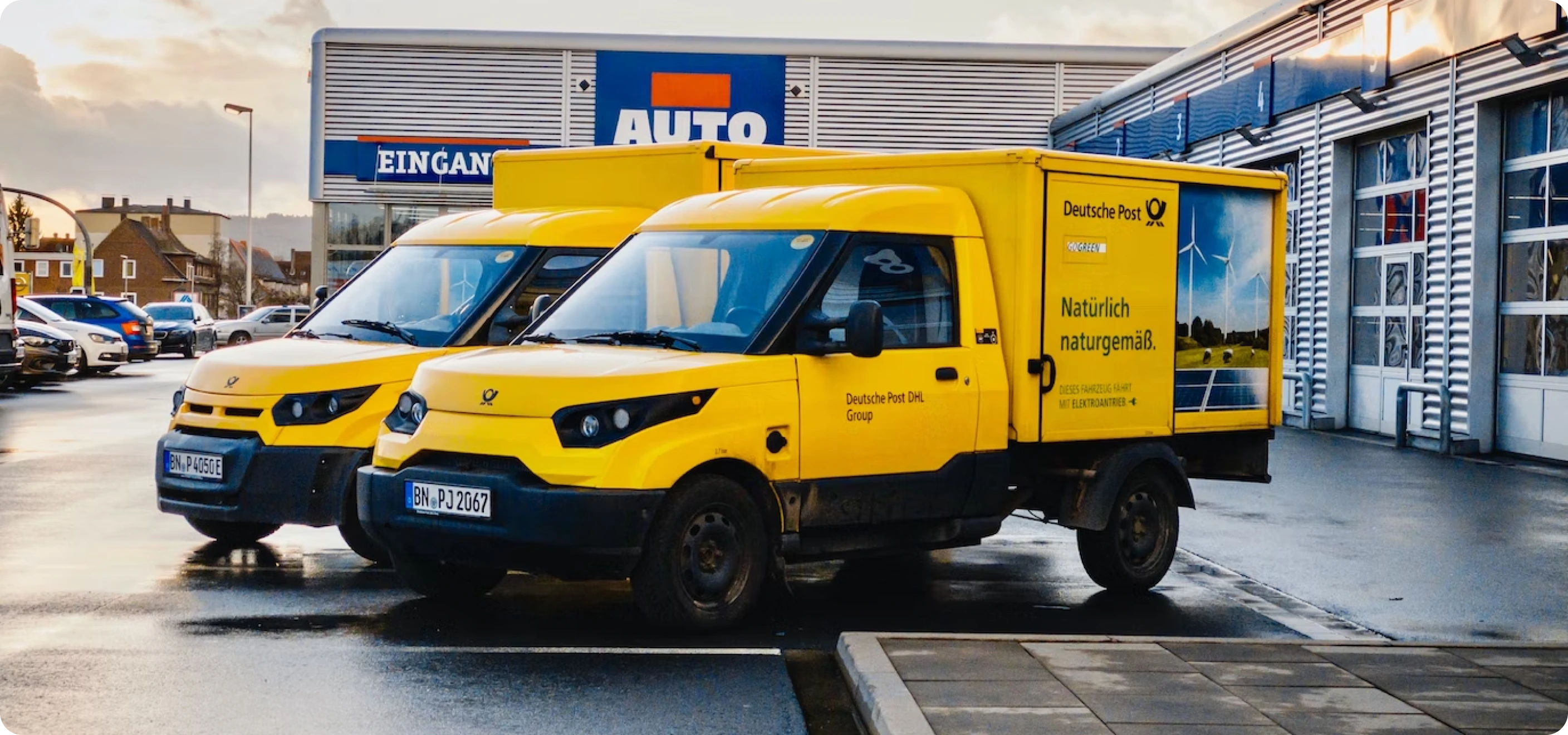 Two yellow Deutsche Post electric delivery vans parked outside an AUTO store.
