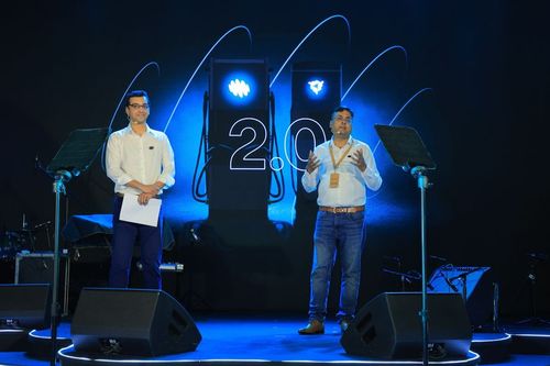 Two speakers on stage at the Exicom 2.0 launch event, with microphones, stage lighting, and a glowing logo backdrop.