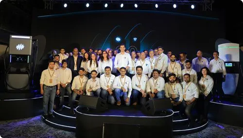 Large group photo of Exicom team on stage in front of branded EV charging units, all wearing white shirts and badges
