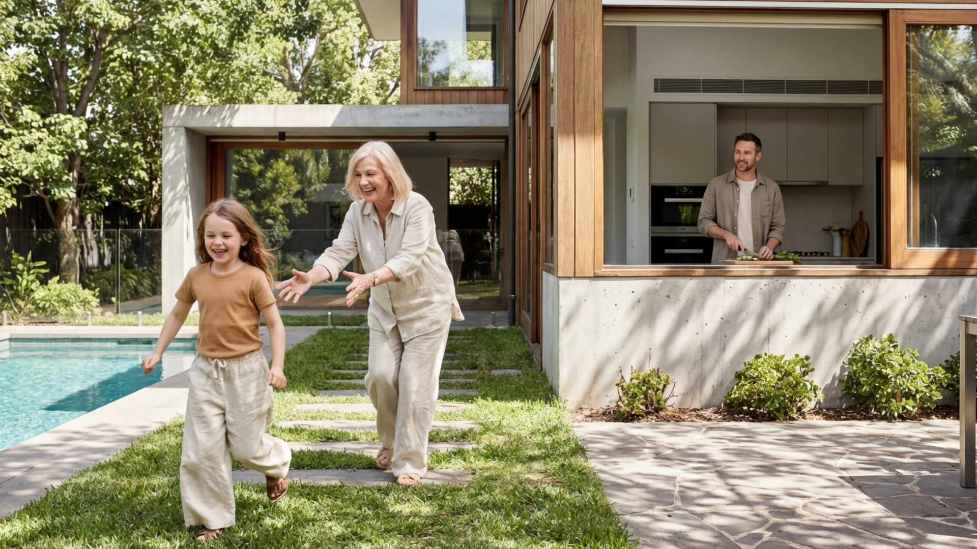 Grandmother reaching to catch smiling girl running on grass beside a pool, with man cooking inside modern house.