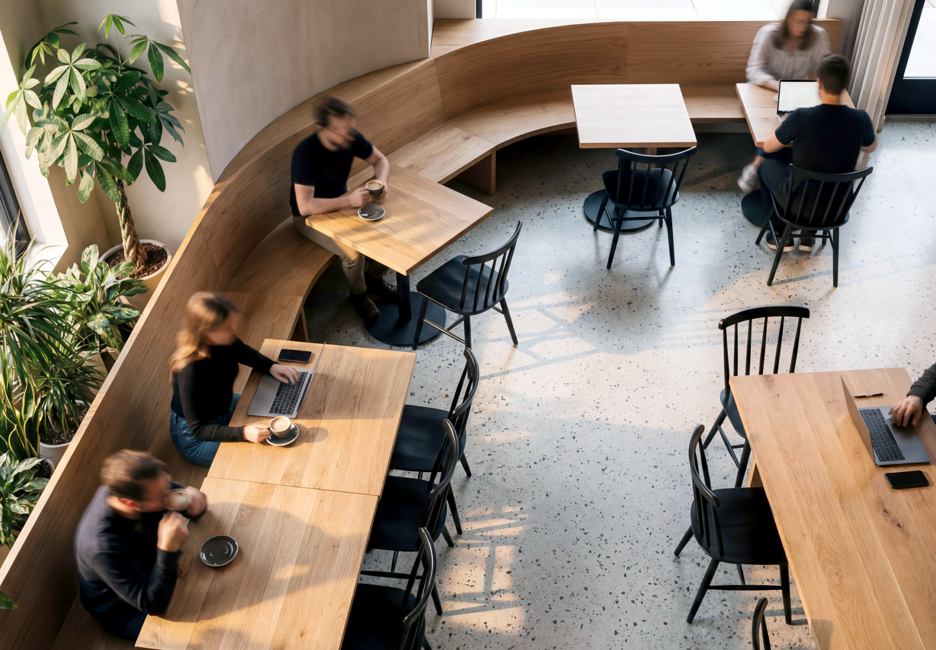 People working on laptops and drinking coffee in a bright cafe with wooden tables and black chairs.