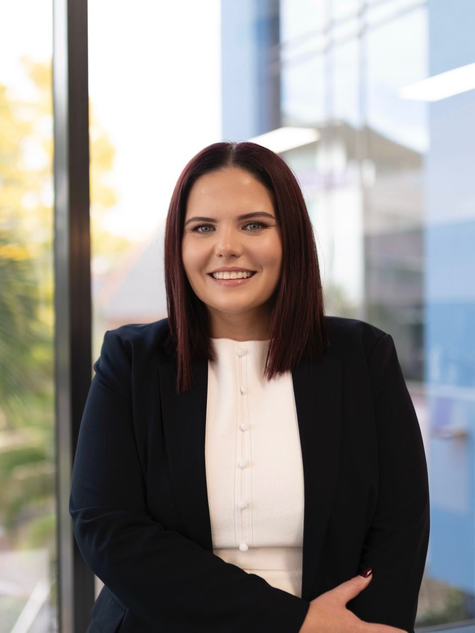 Young professional woman with dark red hair and a black blazer smiling in front of a window.