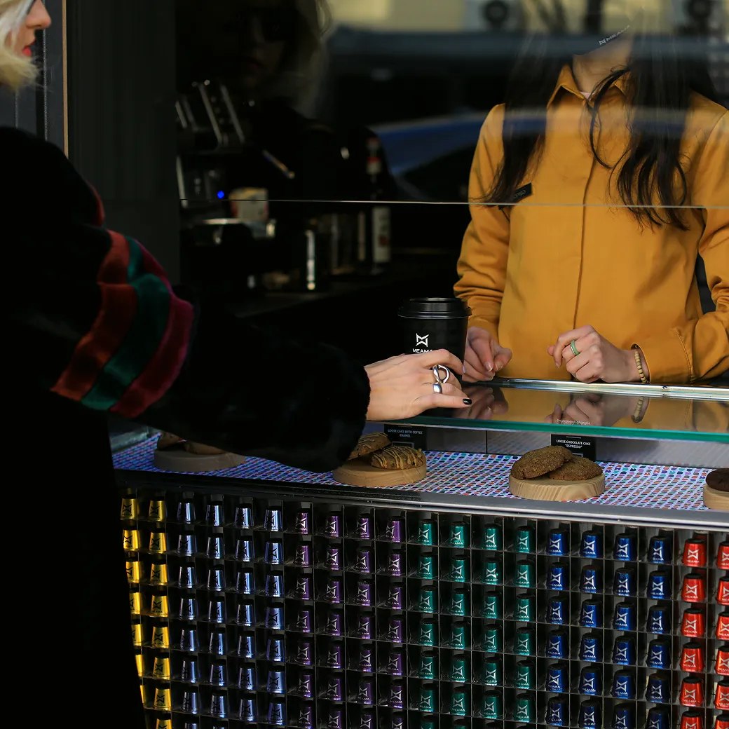 Barista at a Meama Collect coffee shop handing a pink cup to a customer, with coffee capsules displayed on the counter