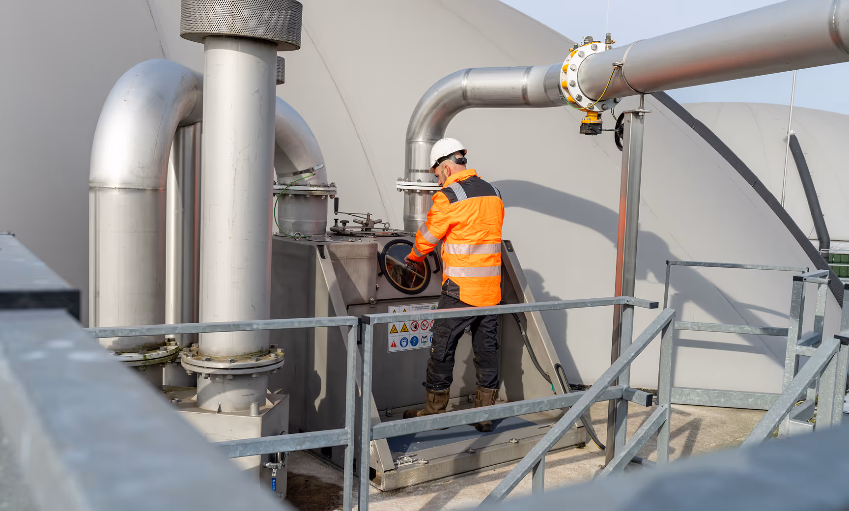 Worker in orange safety jacket and helmet inspecting large industrial pipes and equipment outdoors.