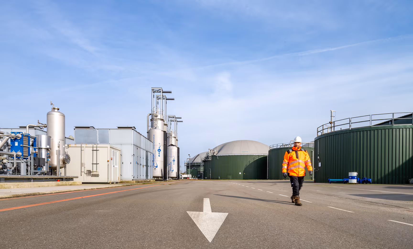 Worker in an orange safety jacket and white helmet walking on a paved road at an industrial plant with large green storage tanks and processing equipment.