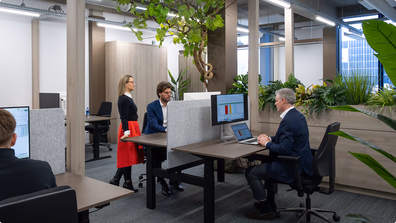 Modern office space with three people working at desks surrounded by plants and natural light.