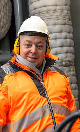 Man in a white safety helmet, yellow ear protectors, and an orange reflective jacket at an industrial site.