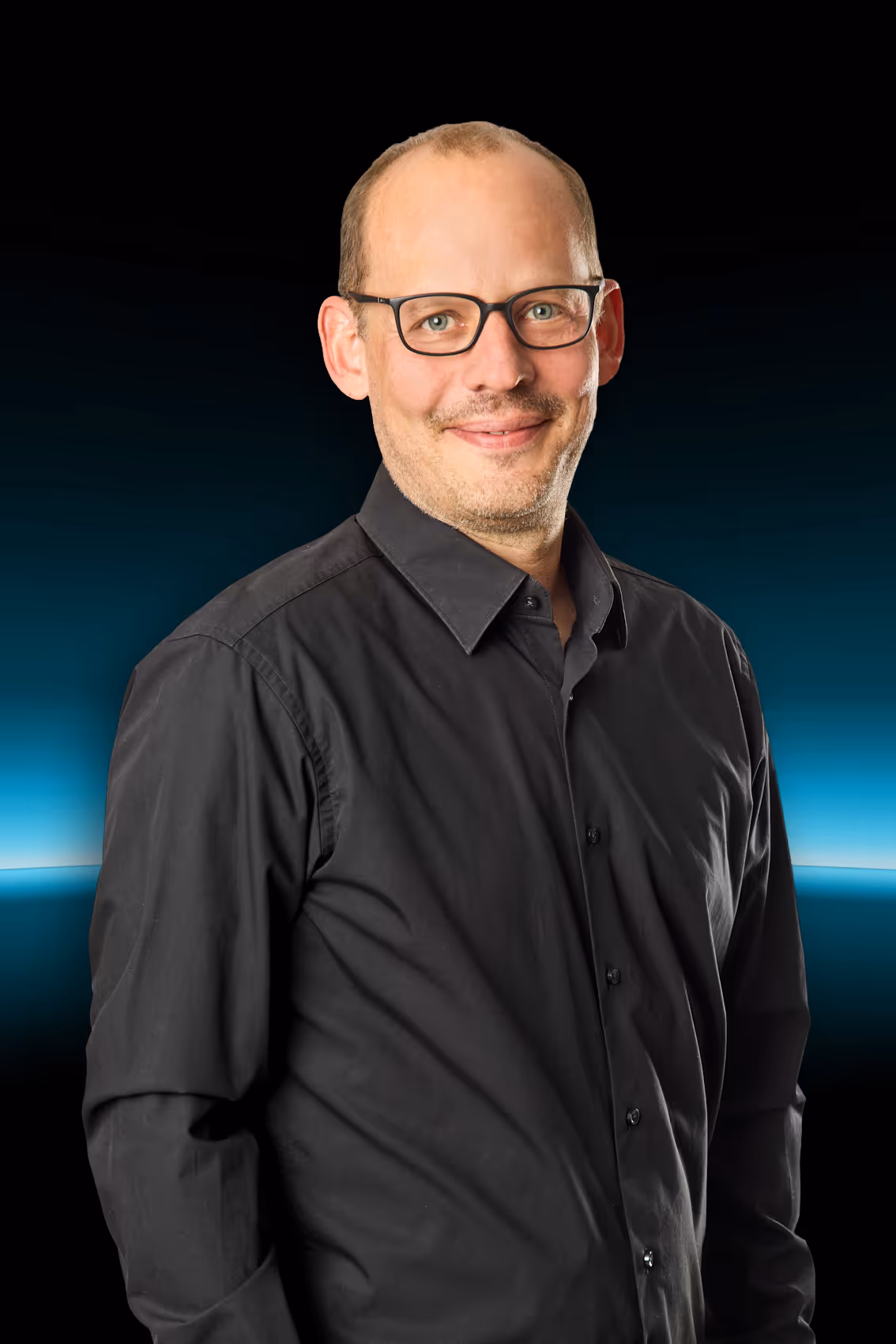 Ein Mann mit Brille und schwarzem Hemd vor einem dunklen Hintergrund mit blauem Lichtschein.