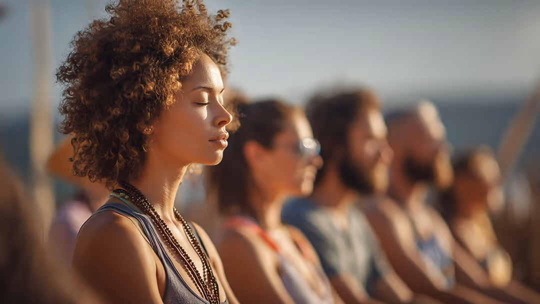 Young woman with curly hair and closed eyes meditating outdoors with a blurred group of people behind her.