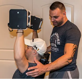 A trainer assists a man lifting dumbbells overhead in a gym setting. The trainer's supportive stance conveys focus and encouragement. Both wear casual workout attire.