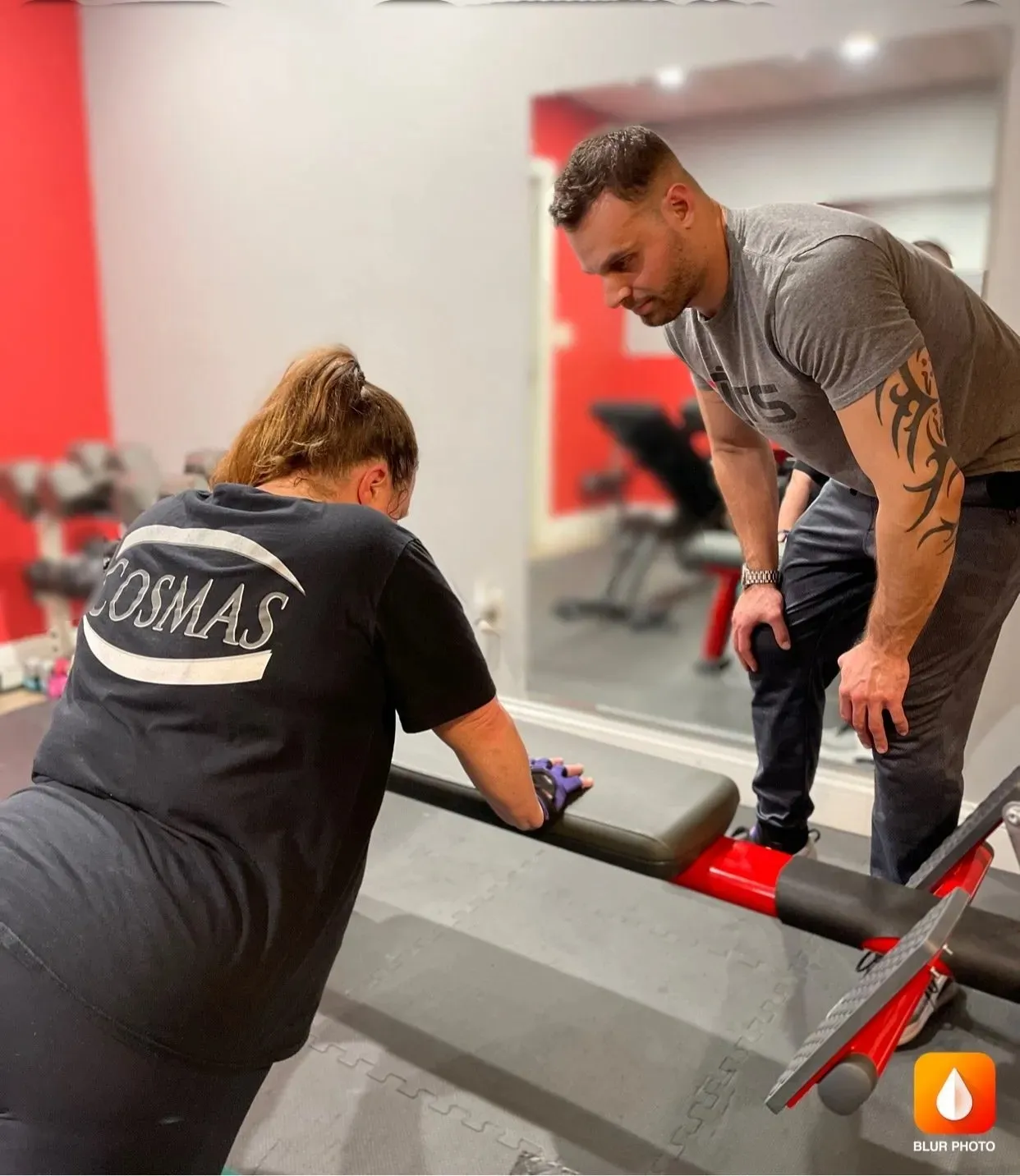 A woman is exercising on a workout bench while a man observes and coaches her in a gym setting.