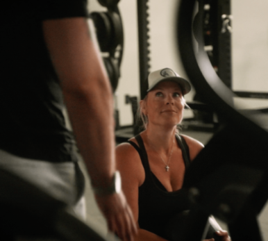 A woman in a gym setting, wearing a cap and tank top, looks up with a smile. She is engaged in a conversation, creating a friendly, motivational vibe.