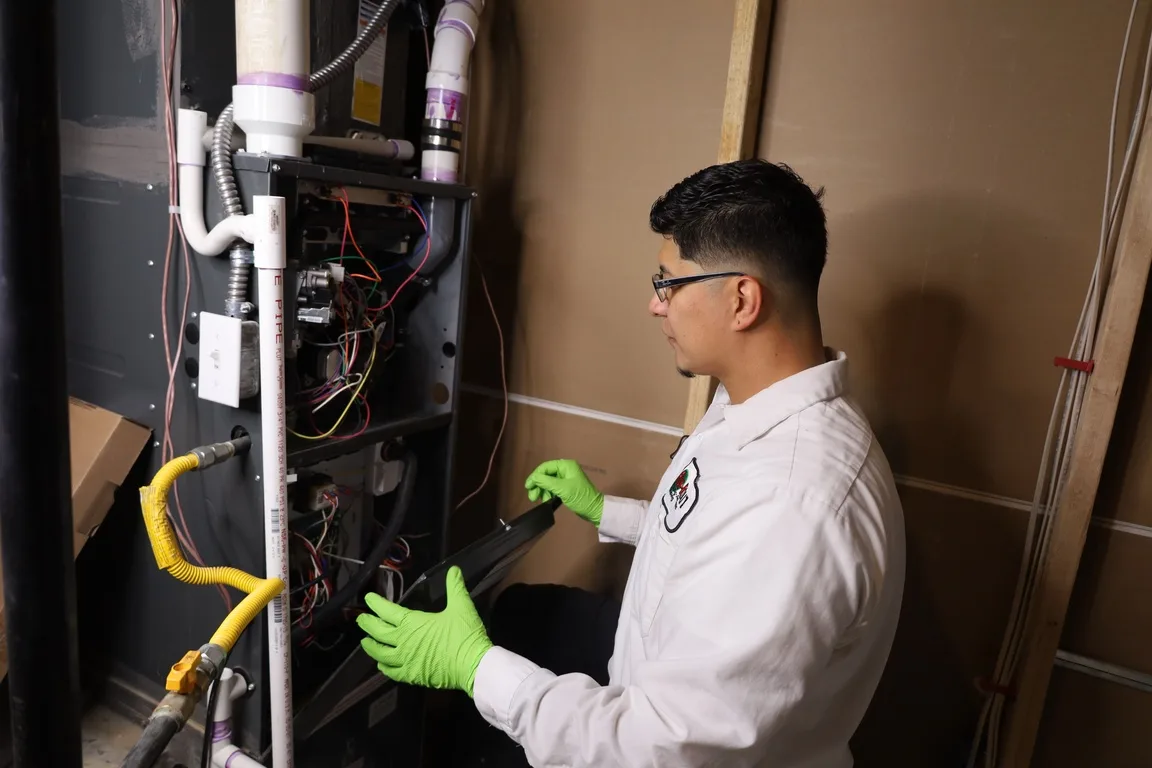 Fix-it 24/7 HVAC technician wearing green work gloves and a white uniform, inspecting an open furnace panel with exposed wiring and PVC venting in a home utility area.