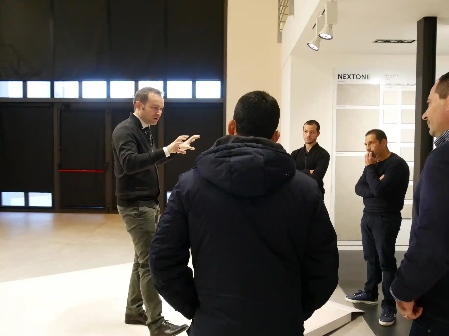 Man gesturing while speaking to a small group of four men indoors near a display board labeled NEXTONE.