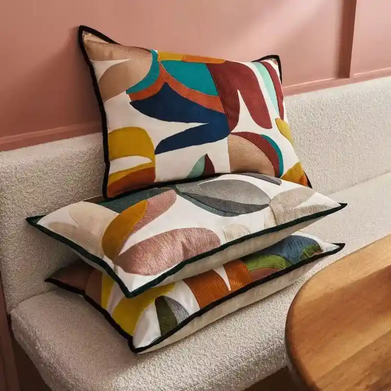 Stack of three decorative pillows with colorful abstract patterns on a textured beige bench seat next to a wooden table and pink wall.