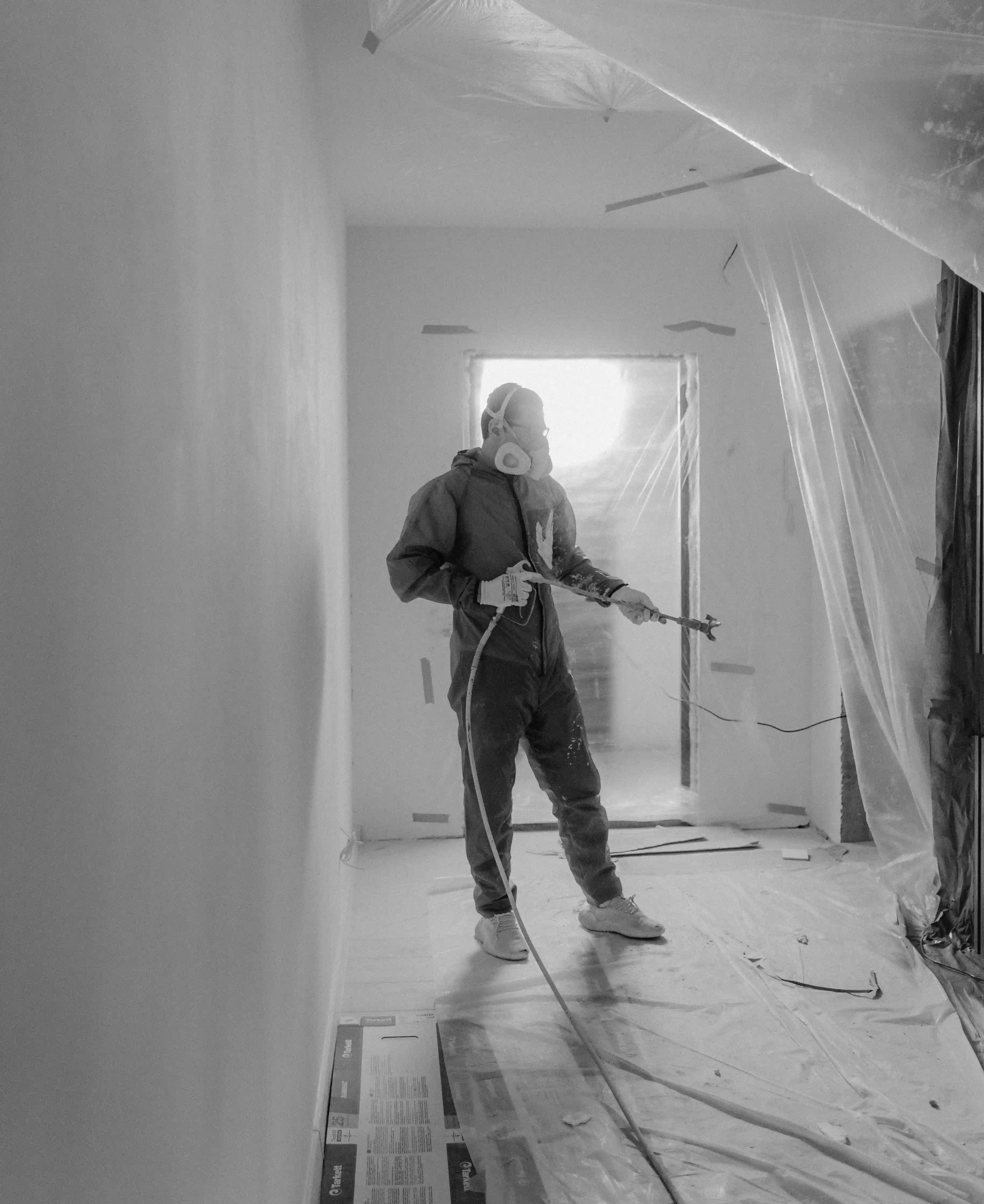 Person wearing protective gear and mask spraying paint or coating inside a room covered with plastic sheets.