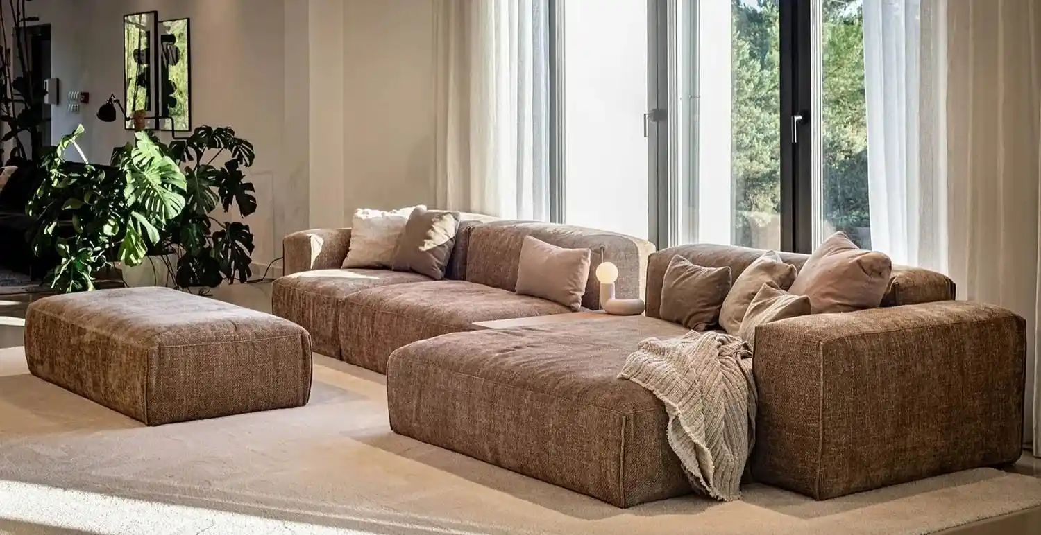 Modern living room with a large brown sectional sofa, assorted cushions, a knitted throw, a small lamp, and a standing plant beside a window with sheer curtains.