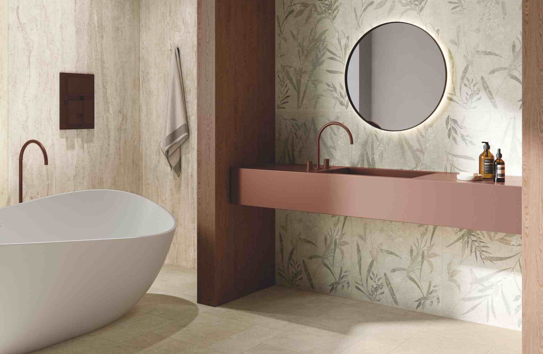 Modern bathroom with white freestanding bathtub, brown wall-mounted sink with copper faucets, circular backlit mirror, and botanical patterned tiles.