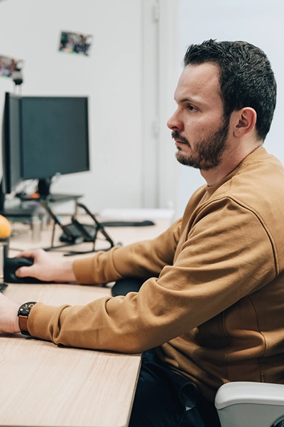 Un homme vêtu d'un pull marron est assis à un bureau, concentré sur un écran d'ordinateur. Environnement de bureau bien rangé et atmosphère calme et concentrée.
