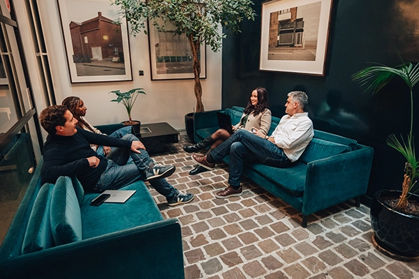Quatre personnes discutent sur des canapés bleu sarcelle dans un salon cosy aux murs sombres. Des œuvres d'art encadrées, des plantes et un sol à motifs de briques créent une atmosphère détendue.
