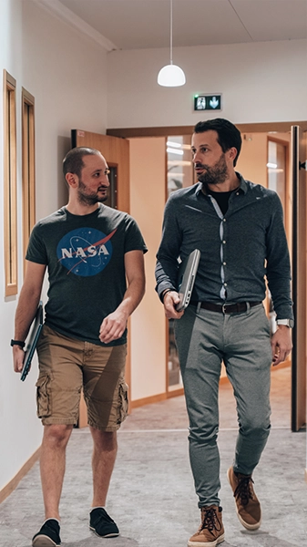 Deux hommes marchent dans un couloir, l'air détendu, chacun avec un ordinateur portable à la main. L'un porte un t-shirt et un short de la NASA, l'autre une chemise boutonnée et un pantalon.